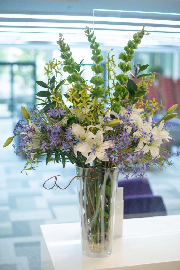 White lilies and blue delphiniums in a tall glass vase in a bright atrium