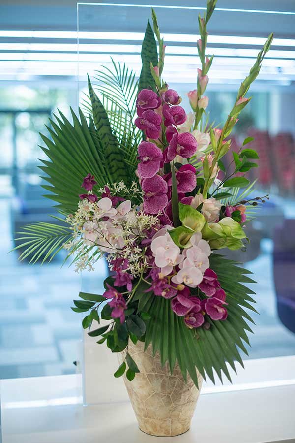 Purple orchids and palm leaves in a tall cream vase by a window