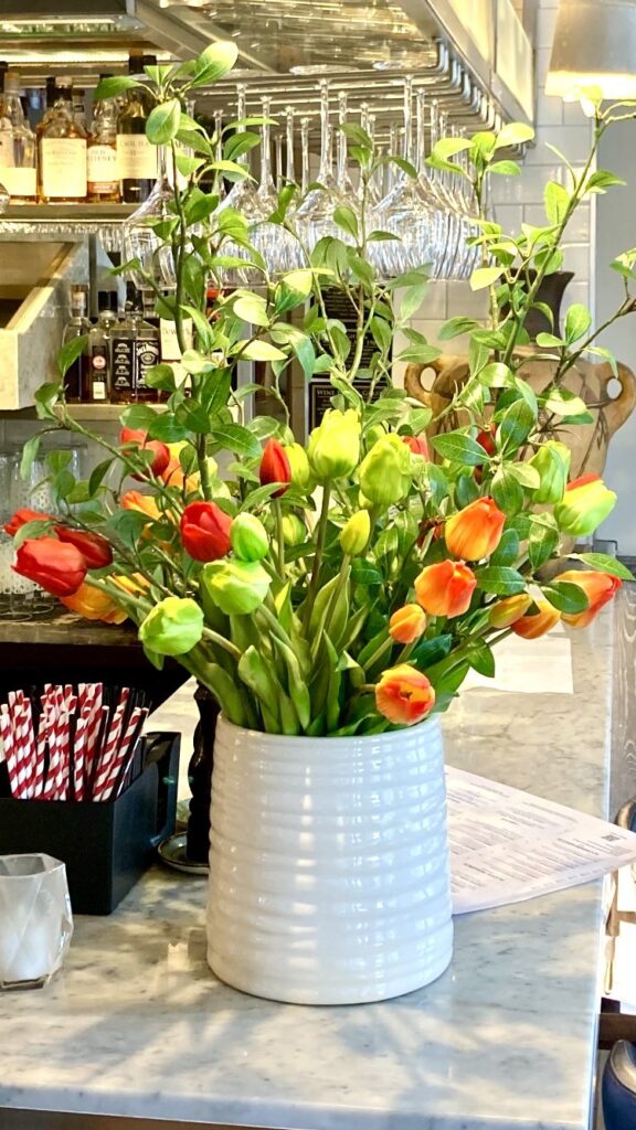 Red and orange tulips with foliage in a white ceramic jug on a marble bar